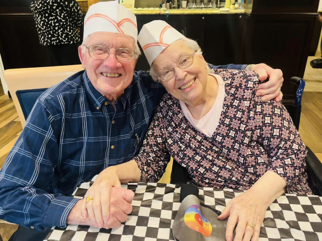 An elderly couple holding hands and smiling while sitting at a table for a happy hour event.