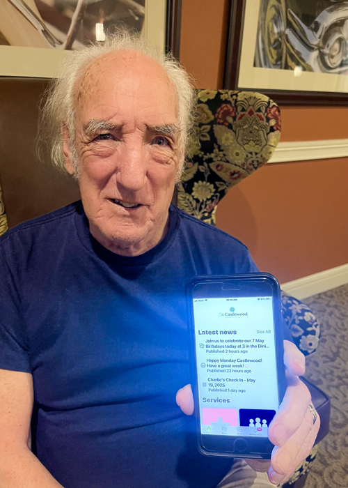 Senior resident seated in a patterned chair, proudly displaying the Arrow app on a smartphone screen during a technology-focused moment.