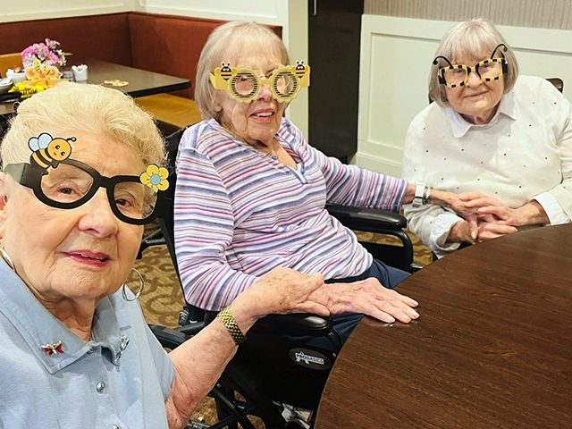 Three senior women wearing playful bee-themed glasses share smiles and hold hands around a table at The Castlewood Senior Living, celebrating community and friendship.