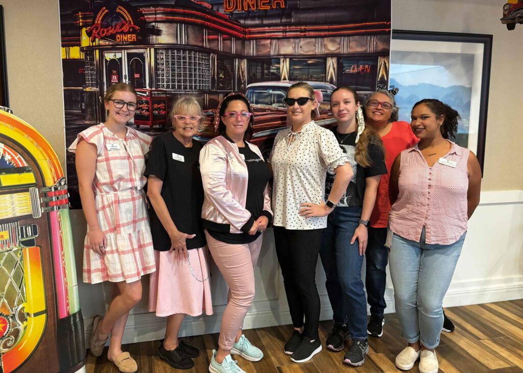 Castlewood Senior Living staff dress in retro attire, posing together in front of a diner backdrop during a themed community event filled with fun and camaraderie.