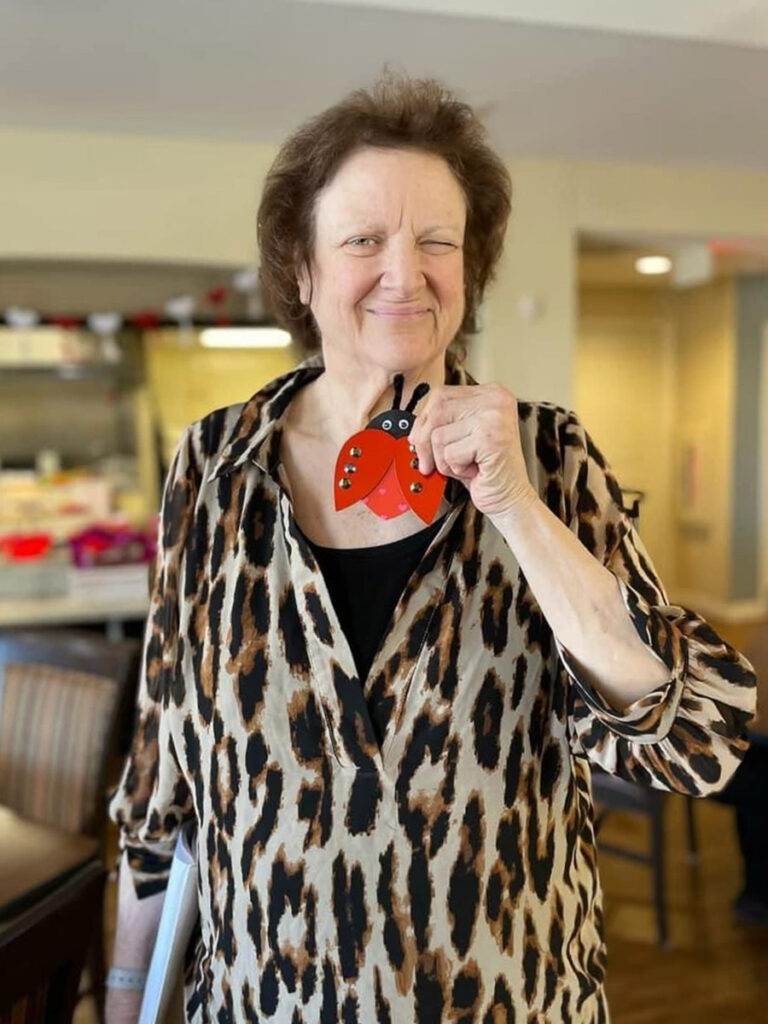 A current photo of Martha in a cheetah print blouse and holding a ladybug paper craft inside the community captures her vibrant personality and artistic flair.
