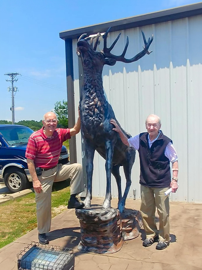 Two elderly men, with smiles on their faces, stand next to a magnificent deer statue, capturing a moment of admiration and friendship.