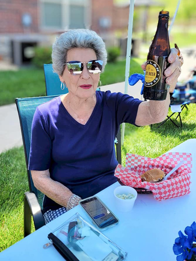 With a beaming smile, an older lady sits in a chair outside, raising a beer bottle in a cheerful toast. On the table is a plate of scrumptious food - a hamburger and coleslaw.