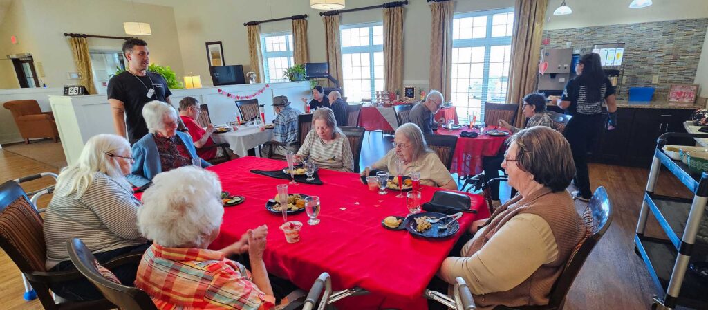 Group of elderly individuals dining together at a table with a red tablecloth in a bright, welcoming room, assisted by line staff.