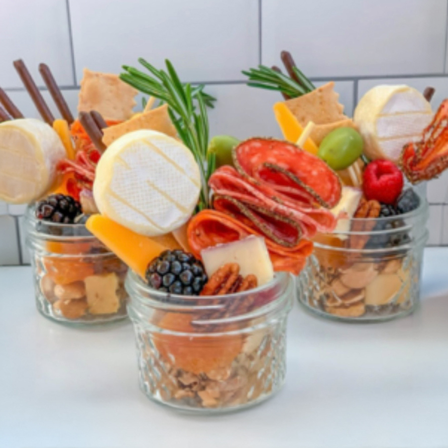 Mini charcuterie cups with cheese, salami, crackers, olives, berries, and rosemary sprigs in glass jars, arranged on a white tiled background.