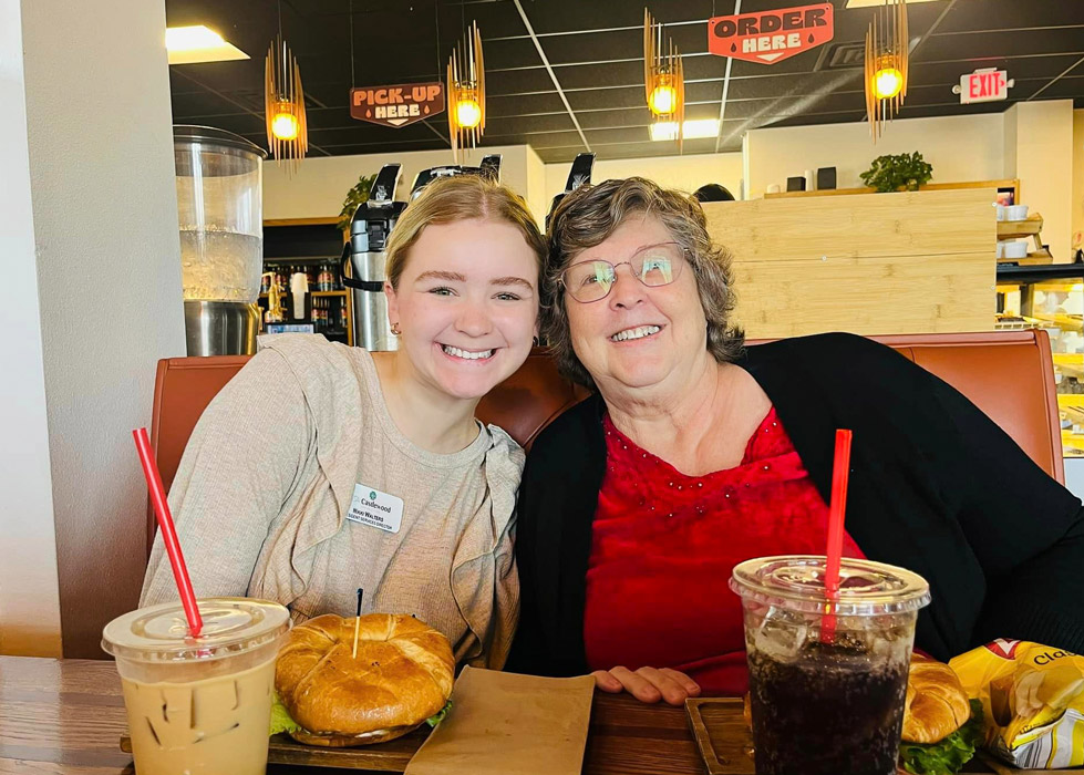 A senior living employee and resident enjoy a meal together at Mudhouse Coffee. They are seated side by side, smiling warmly, with sandwiches and drinks in front of them, capturing a moment of connection and companionship.