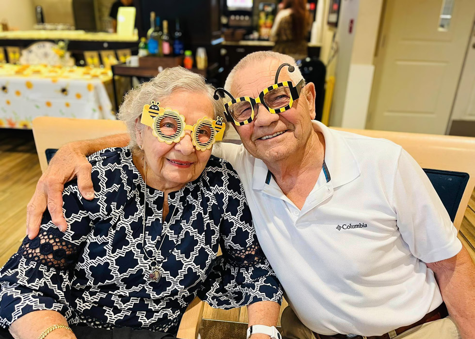 A senior couple, dressed in playful bee-themed glasses, sit close together and smile warmly at the camera during a themed event, likely "Don't Step on a Bee Day."