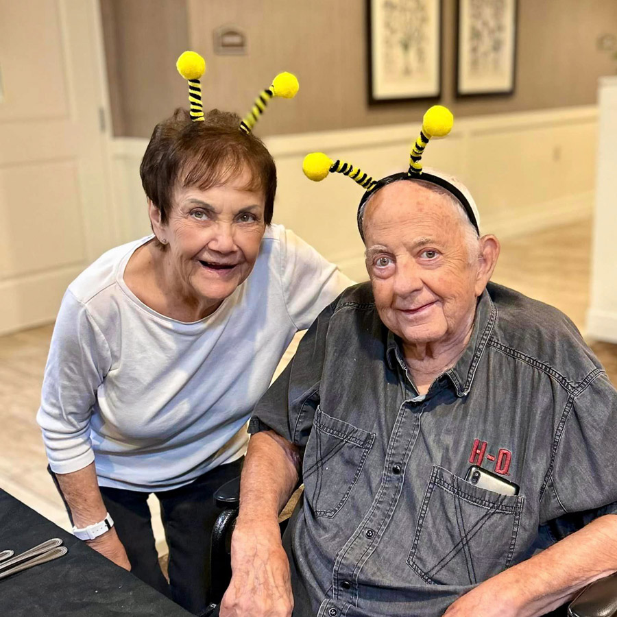 A senior couple, both wearing bee-themed headbands with yellow antennae, smile together during a "Don't Step on a Bee Day" event. The cheerful accessories add a fun and festive touch to their day.