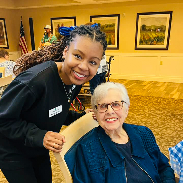 A senior living employee and resident share a joyful moment together. The employee leans in beside the seated resident, both smiling warmly. The setting appears to be a lively gathering or event, with other residents and a event visible in the background.