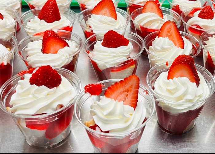 Individual strawberry shortcake desserts in clear cups, topped with whipped cream and a fresh strawberry slice, arranged in rows on a serving tray.