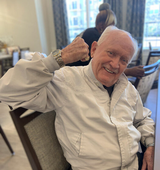 Smiling senior resident in a white jacket raises his arm in a playful flexing gesture while seated in a senior living dining area, exuding confidence and joy.