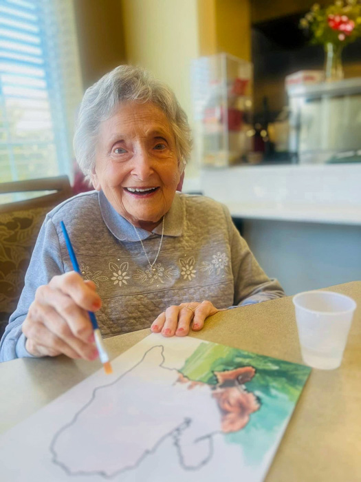 Senior resident happily painting at a table, holding a paintbrush while working on a colorful art project, enjoying creative time in a bright and cheerful senior living community.