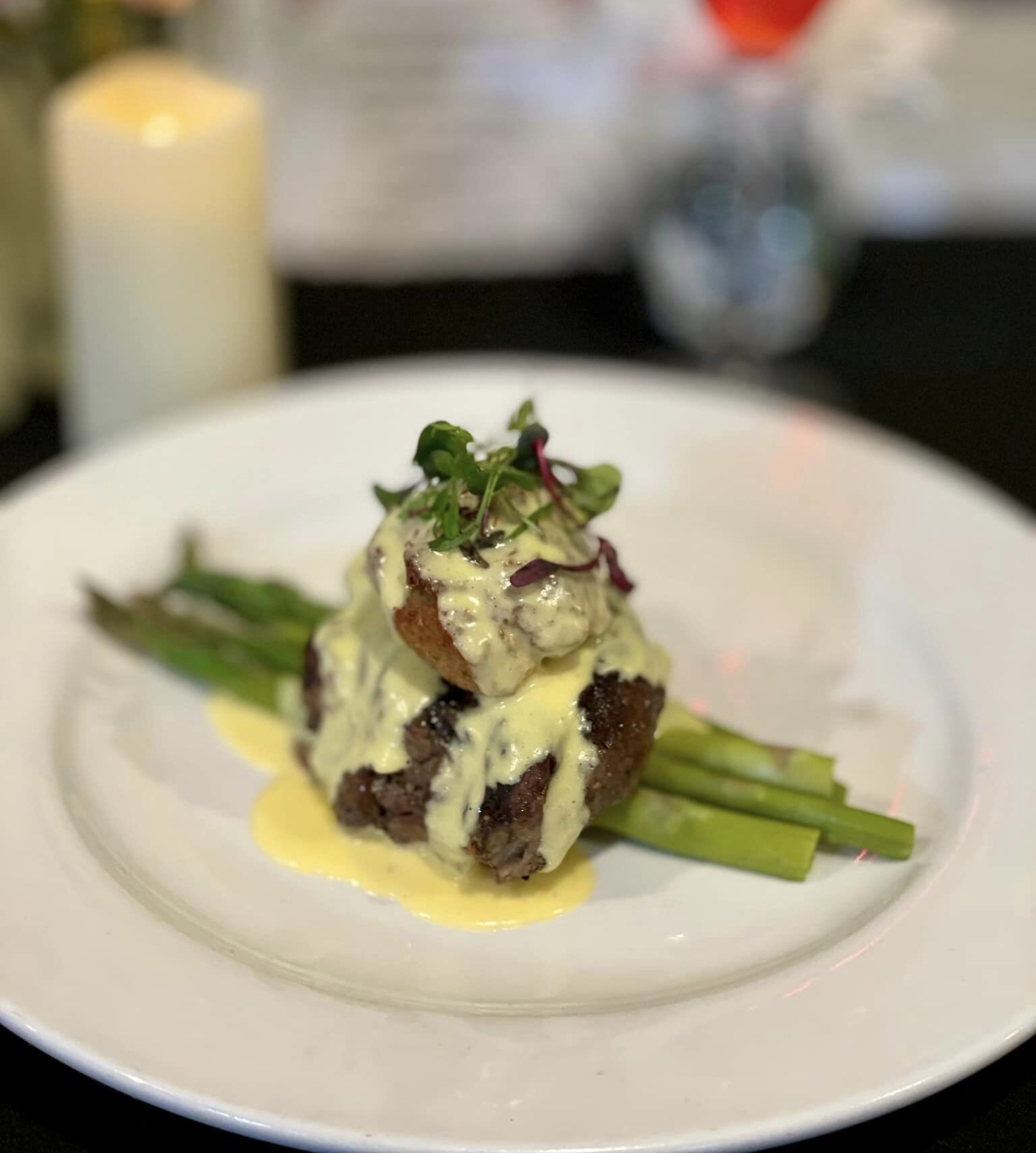 Plated gourmet steak with asparagus, topped with creamy hollandaise sauce and garnished with microgreens, set against a blurred background of a candle, flowers, and drink.