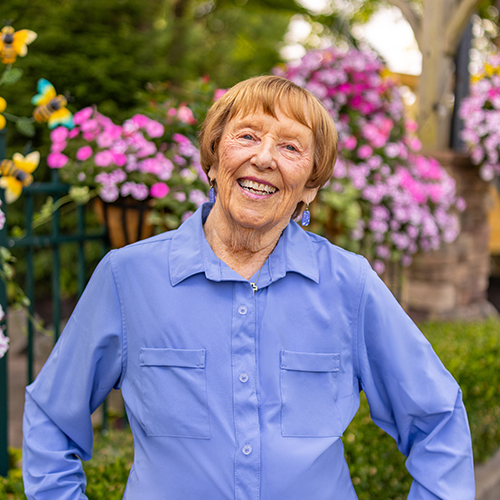 A senior resident wearing a light blue shirt smiles brightly while standing in front of vibrant pink flowers in a beautifully landscaped outdoor setting.
