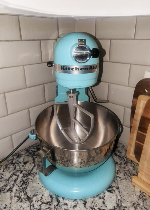 Aqua blue KitchenAid stand mixer with a stainless steel bowl and paddle attachment on a granite countertop, placed in a tiled kitchen corner.