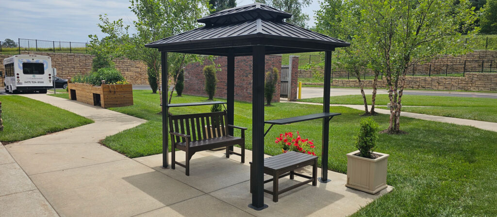 Gazebo with bench and flowers on a sunny day at The Castlewood Senior Living, surrounded by trees, raised garden beds, and a community shuttle.