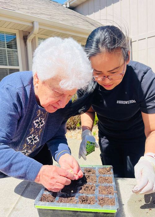 Elderly woman and team member planting seeds in soil starter tray during a sunny outdoor gardening activity at The Castlewood Senior Living.