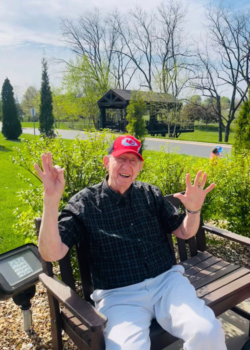 Senior man in red Kansas City Chiefs hat happily waving while sitting on a bench surrounded by spring greenery.