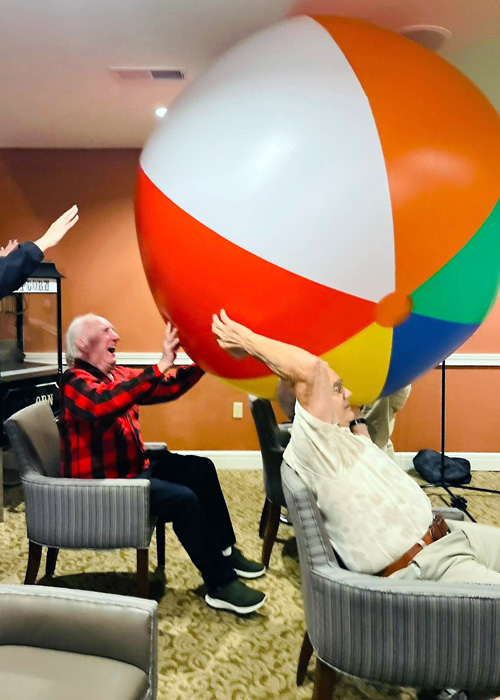 Senior residents laugh and play with a giant beach ball during a lively indoor game, staying active and engaged in a fun-filled community wellness event.