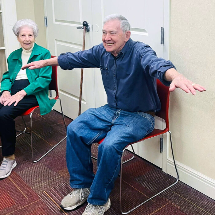 Senior resident stretches with arms out during a seated wellness session, smiling brightly while participating in a group movement activity alongside peers.
