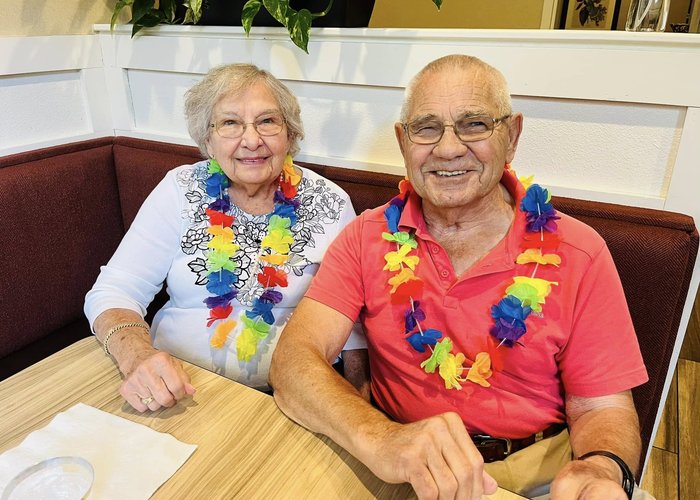 A smiling senior resident couple wearing colorful leis sits side by side in a booth at The Castlewood Senior Living.