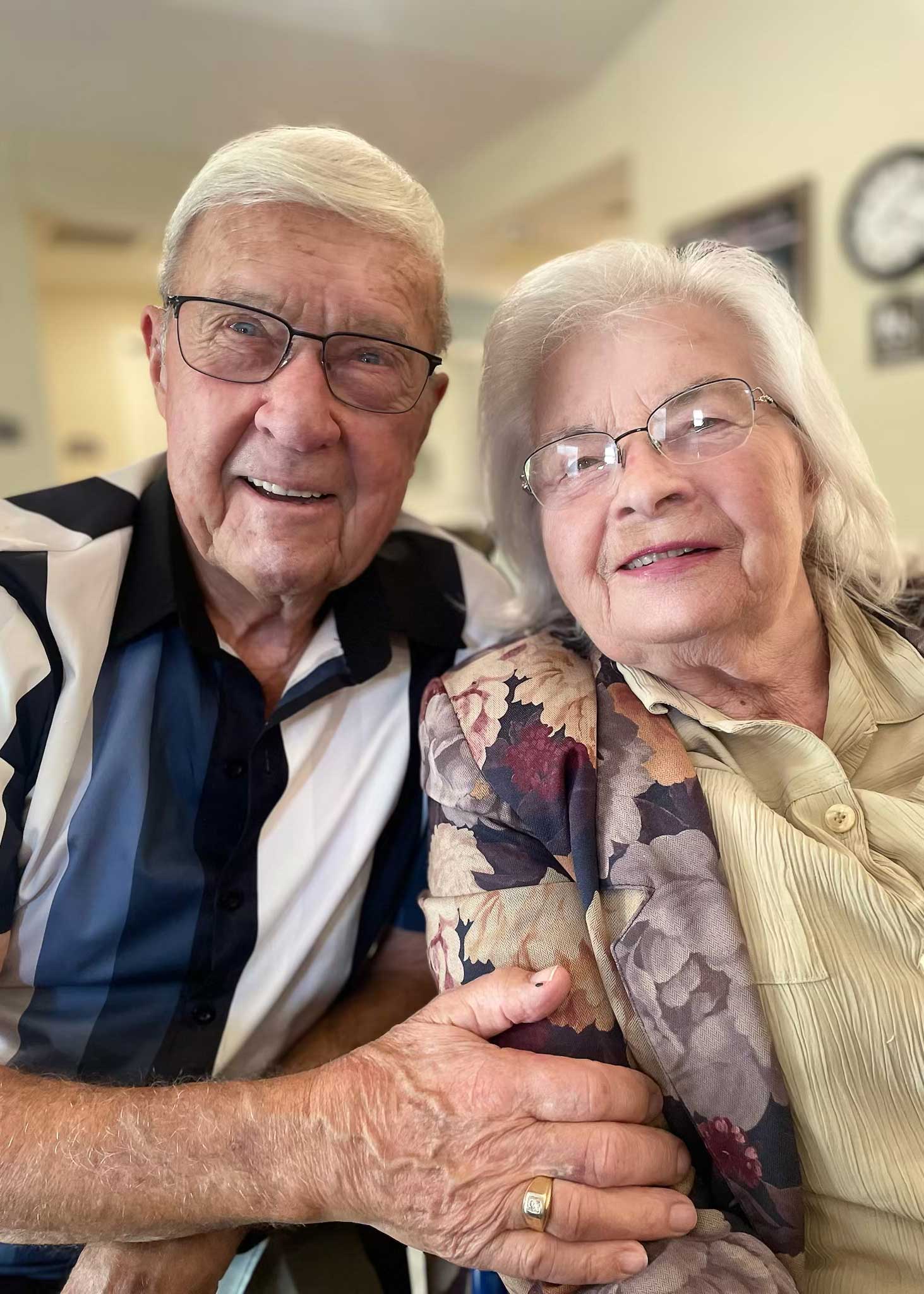 A smiling senior couple sits closely together, both wearing glasses, sharing a warm embrace and joyful moment inside their welcoming senior living community.