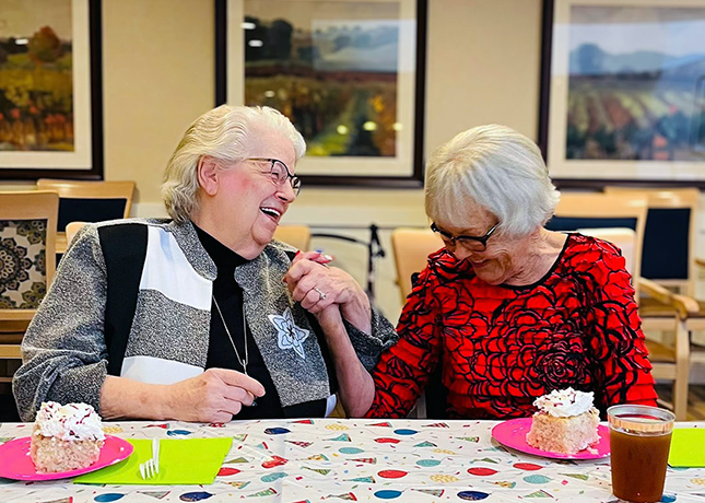 Two senior women clasp hands, laughing, at a decorated table with slices of birthday cake.