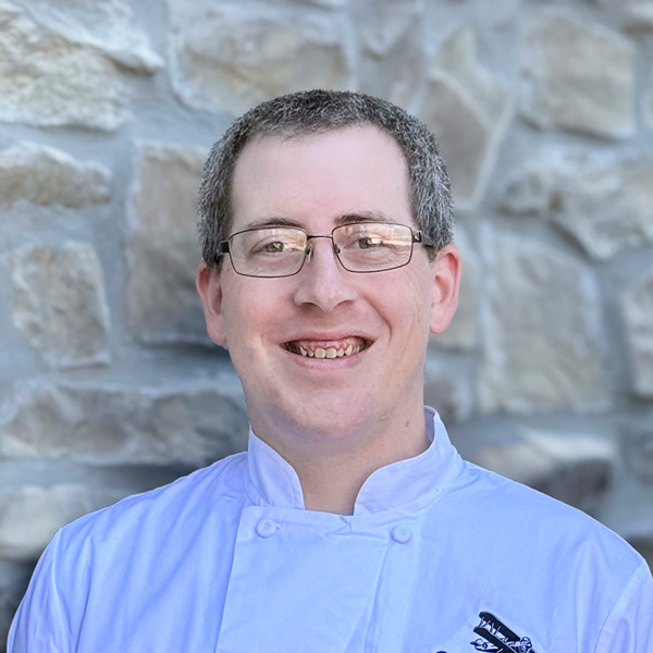 Cody Keel, Culinary Director at The Castlewood Senior Living, smiling in a white chef coat while standing in front of a light stone wall.