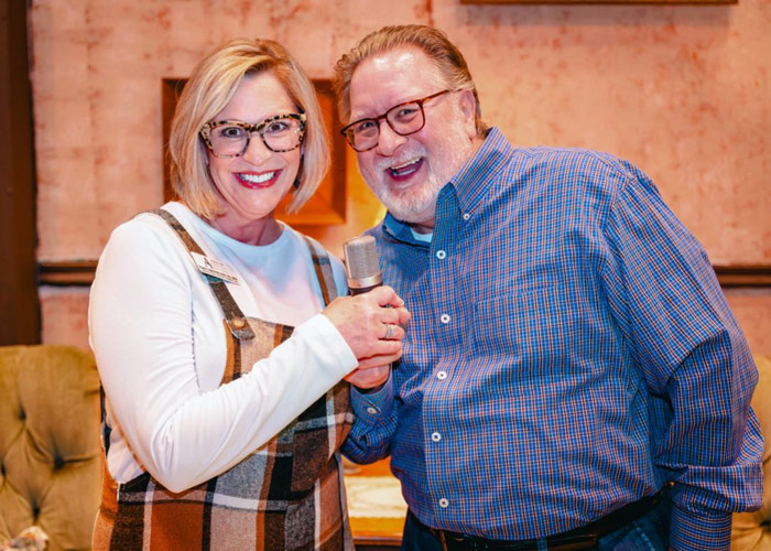 Elizabeth and Charlie, smiling hosts of Thoughtful Connections: A Memory Care Podcast, stand close together holding a microphone in a warmly lit studio at Cottey College.