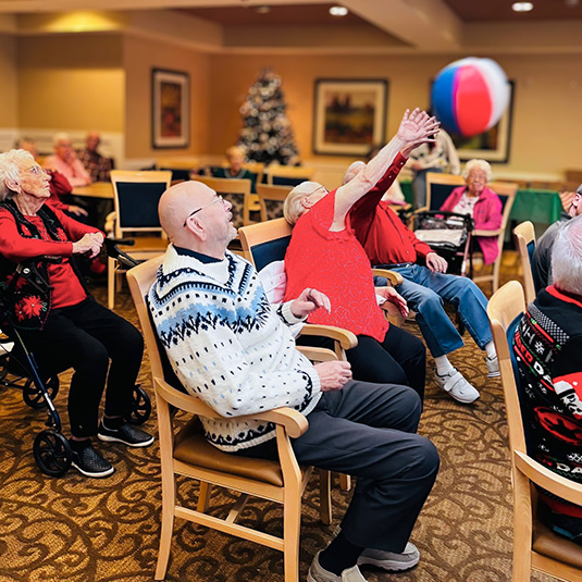 Seniors reach up excitedly, playing chair beachball volleyball at The Castlewood Senior Living.