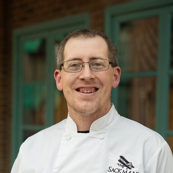 Cody Keel, Culinary Director at The Castlewood Senior Living, smiling in a professional headshot, wearing glasses and a white chef coat, with a softly blurred brick and window background.