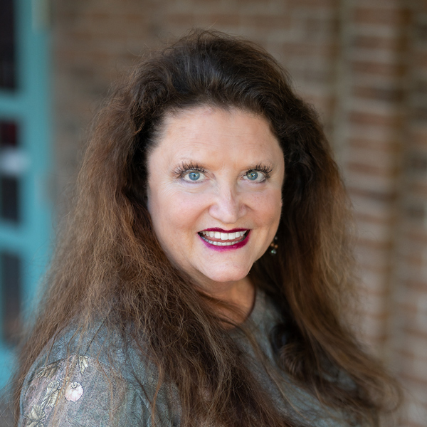 Phyllis Wiley, Senior Living Director at The Castlewood Senior Living, smiling in a professional outdoor headshot with long dark wavy hair, bright lipstick, and a softly blurred brick background.