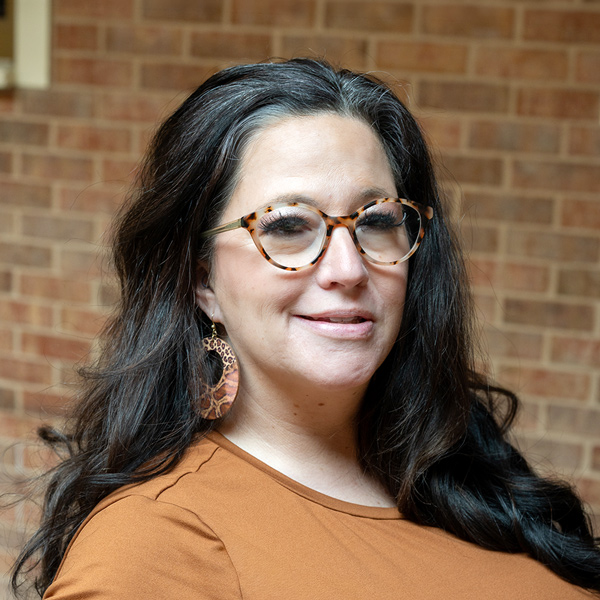 Sara Smith, Resident Services Director at The Castlewood Senior Living, in a professional outdoor headshot with long dark hair, tortoiseshell glasses, large patterned earrings, a brown top, and a softly blurred brick background.