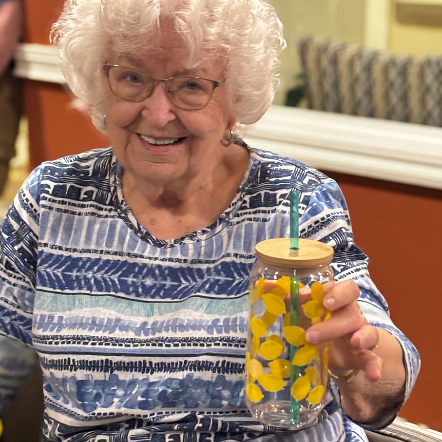 A senior woman smiles, holding up a floral drinking glass with straw, at The Castlewood Senior Living.