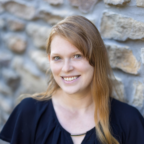 Meghan Reynolds, Business Office Director at The Castlewood Senior Living, smiling in a professional headshot with long strawberry blonde hair, a black blouse, and a softly blurred stone wall background.