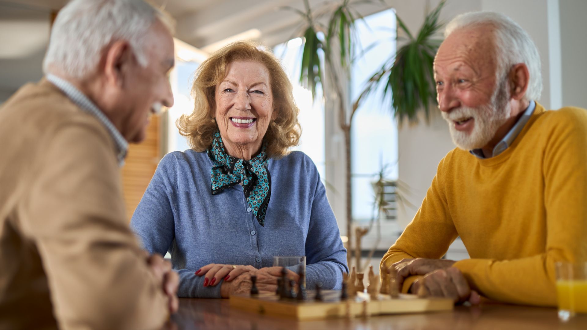 Three older adults smiling and laughing together while playing chess at a table indoors.