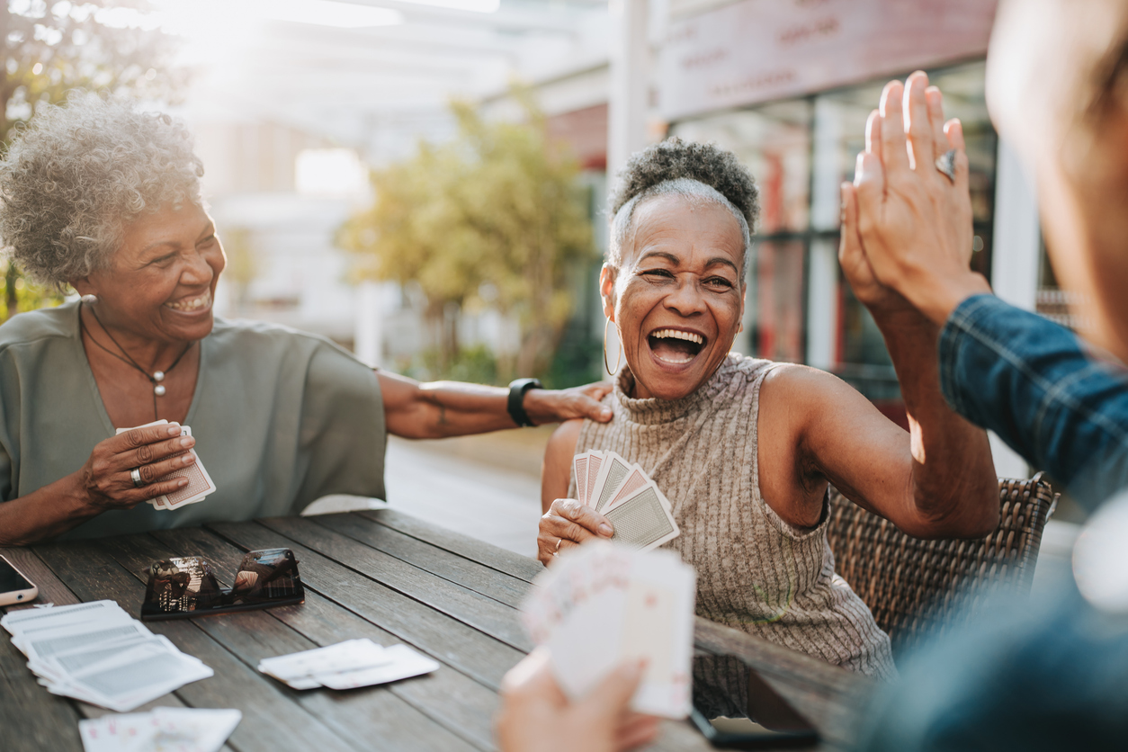 Smiling senior women laughing and playing cards outdoors, sharing a high five at a wooden table