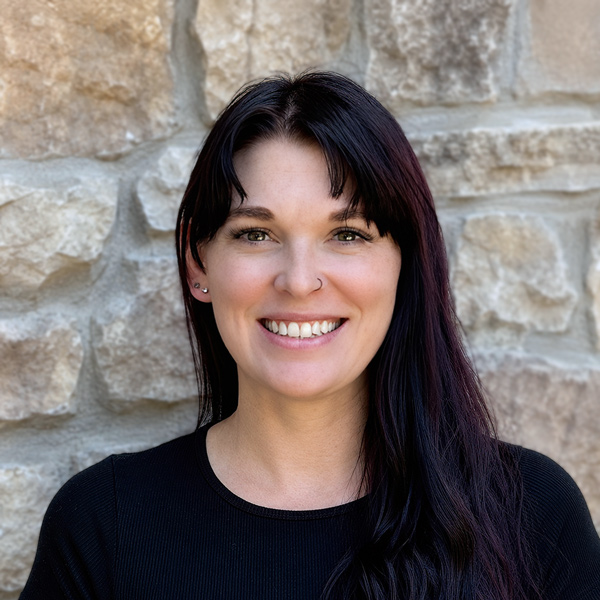 Meghan “Meg” Reynolds, Business Office Director at The Castlewood Senior Living, smiling in a professional headshot with long dark hair, a black top, and a light stone wall background.