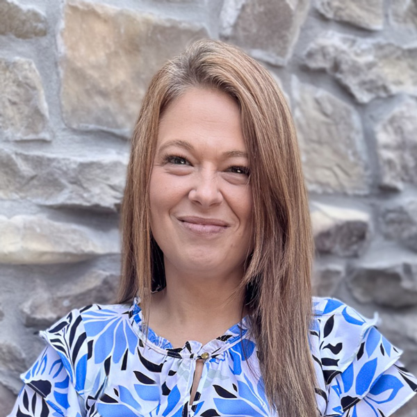 Sarah Umbenhower, Memory Care Director at The Castlewood Senior Living, smiling in a professional headshot wearing a blue floral blouse before a stone wall.